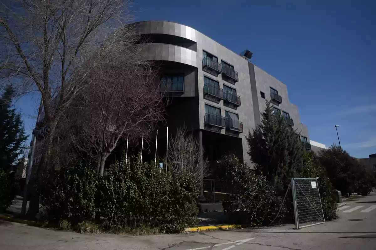 Edificio moderno de varios pisos con fachada de tonos oscuros rodeado de árboles y arbustos bajo un cielo despejado Edificio moderno de varios pisos con fachada de tonos oscuros rodeado de árboles y arbustos bajo un cielo despejado