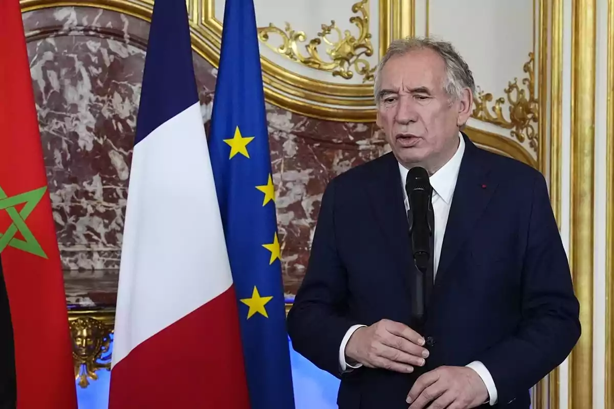 Hombre de traje azul hablando frente a un micrófono con las banderas de Marruecos, Francia y la Unión Europea al fondo en una sala decorada elegantemente