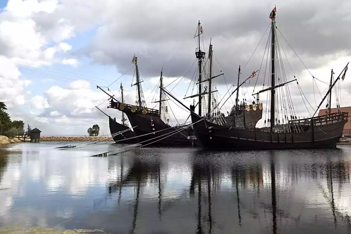 Tres carabelas de madera están ancladas en un puerto tranquilo bajo un cielo nublado, reflejándose en el agua.
