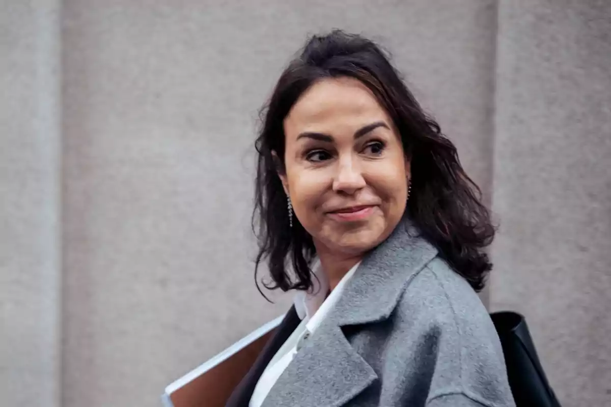 Mujer de cabello oscuro con abrigo gris sonriendo y mirando hacia un lado mientras camina frente a una pared clara Mujer de cabello oscuro con abrigo gris sonriendo y mirando hacia un lado mientras camina frente a una pared clara