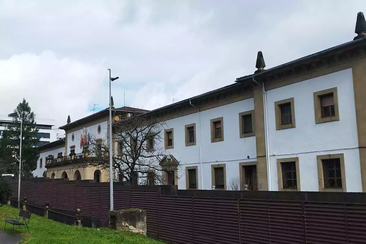 Edificio antiguo de fachada blanca con detalles en piedra y ventanas enmarcadas, rodeado por una cerca y un área verde, bajo un cielo nublado. Edificio antiguo de fachada blanca con detalles en piedra y ventanas enmarcadas, rodeado por una cerca y un área verde, bajo un cielo nublado.