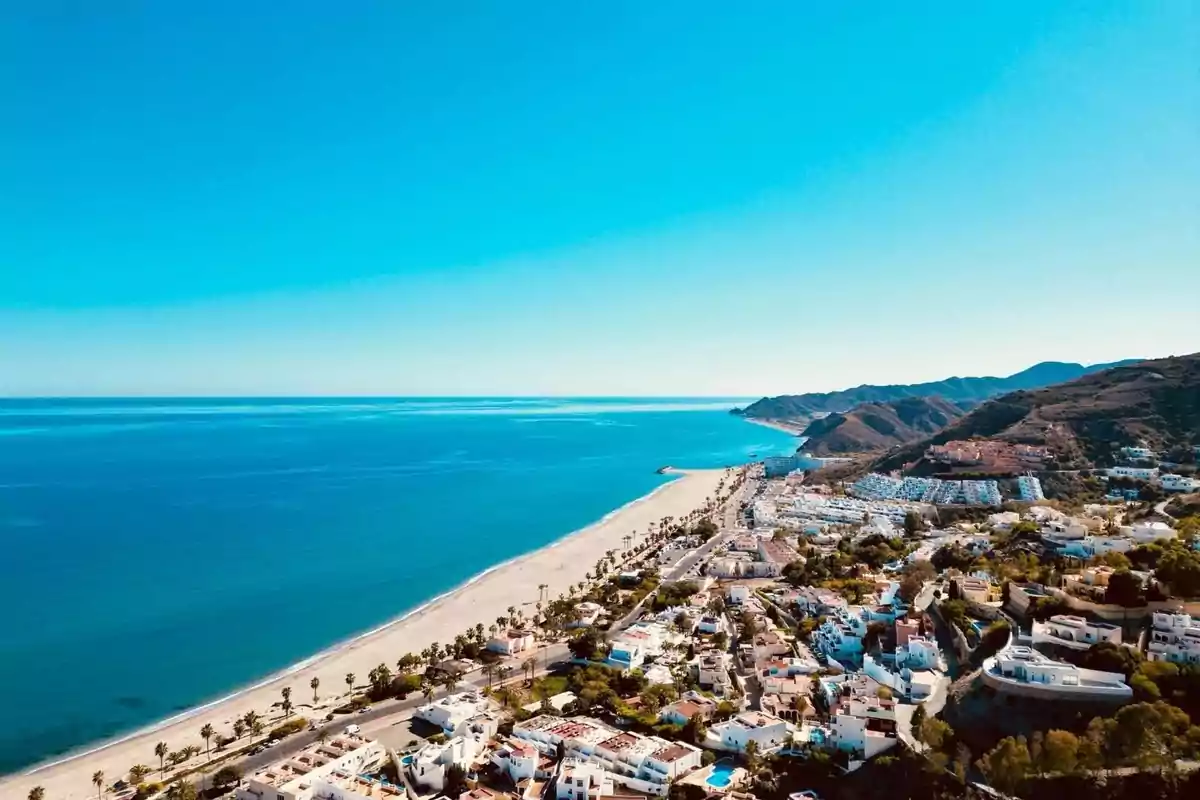 Vista aérea de una playa de arena clara junto a un pueblo con casas blancas y montañas al fondo bajo un cielo despejado