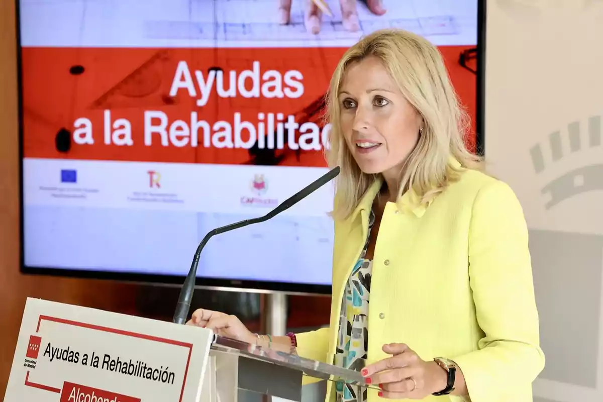 Mujer rubia con chaqueta amarilla hablando en un atril durante una presentación sobre ayudas a la rehabilitación con una pantalla de fondo que muestra el mismo tema Mujer rubia con chaqueta amarilla hablando en un atril durante una presentación sobre ayudas a la rehabilitación con una pantalla de fondo que muestra el mismo tema