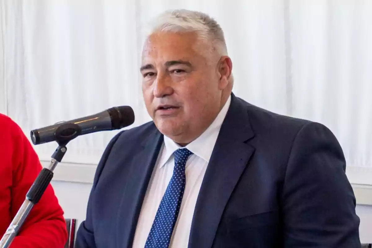 Hombre de cabello canoso y corto usando traje azul y corbata azul con puntos blancos hablando frente a un micrófono Hombre de cabello canoso y corto usando traje azul y corbata azul con puntos blancos hablando frente a un micrófono