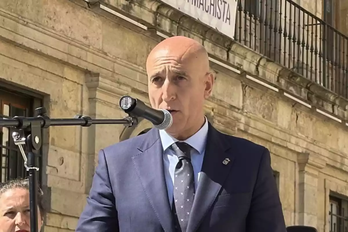 Hombre calvo con traje azul hablando frente a un micrófono en un evento al aire libre con edificio de piedra de fondo Hombre calvo con traje azul hablando frente a un micrófono en un evento al aire libre con edificio de piedra de fondo