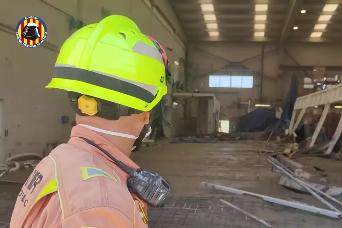Bombero con casco amarillo y uniforme naranja observa el interior de una nave industrial con escombros y daños estructurales