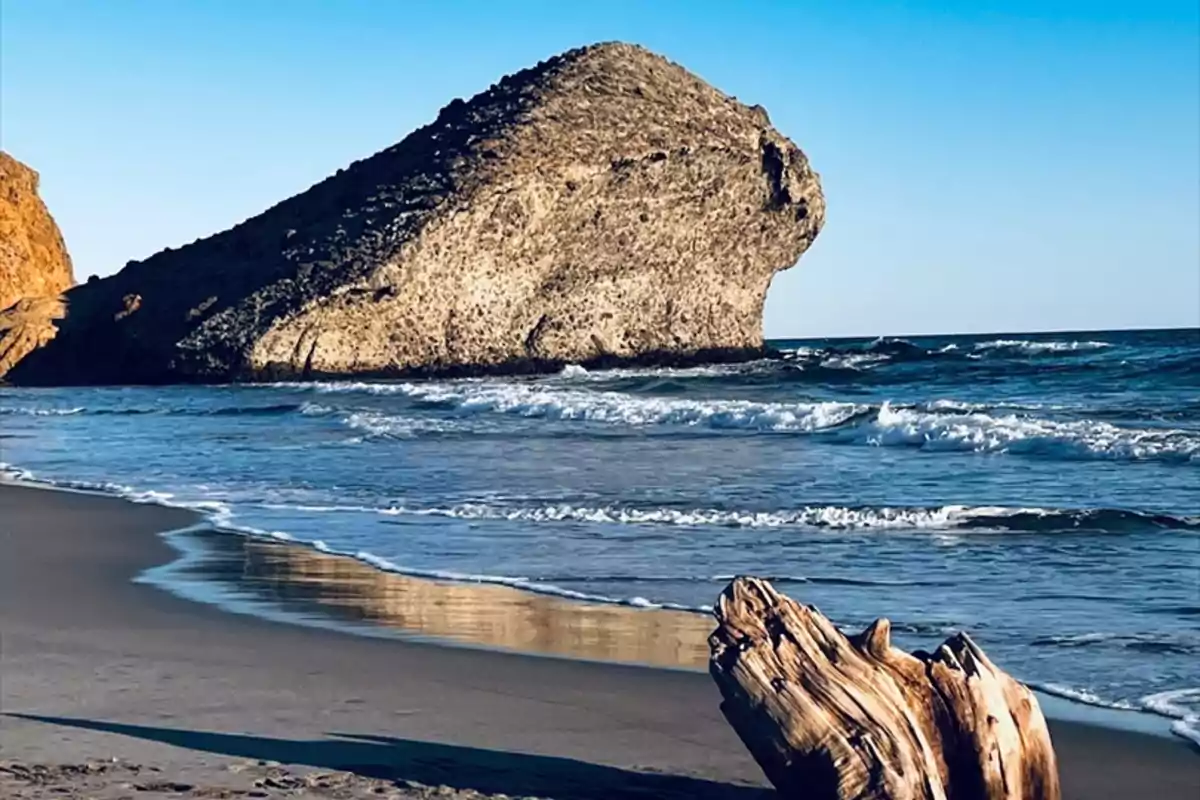 Playa con una gran roca junto al mar y un tronco en la arena bajo un cielo despejado Playa con una gran roca junto al mar y un tronco en la arena bajo un cielo despejado