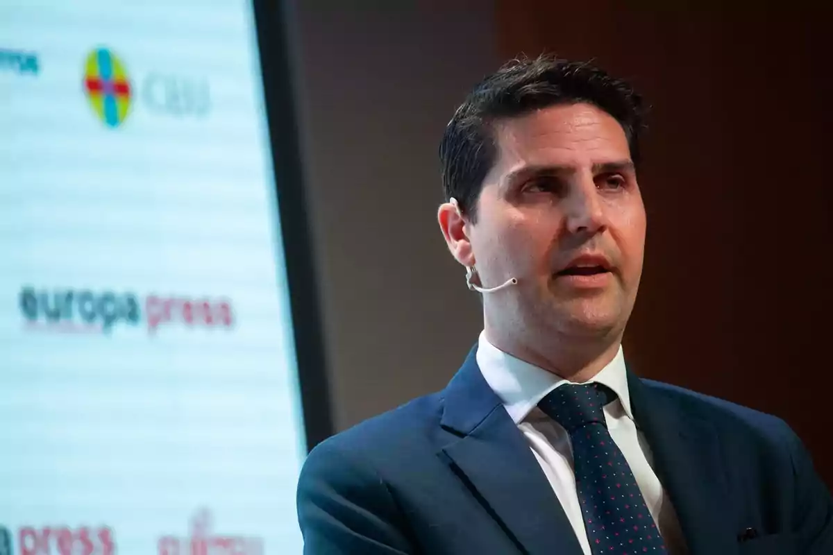 Hombre de cabello oscuro con traje azul y corbata hablando en un evento con micrófono de diadema y una pantalla de fondo con logotipos.