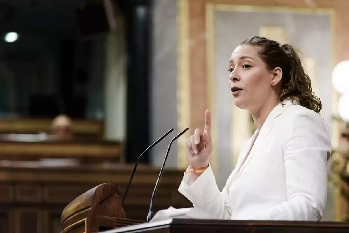 Mujer con chaqueta blanca hablando y gesticulando en un podio dentro de un recinto parlamentario Mujer con chaqueta blanca hablando y gesticulando en un podio dentro de un recinto parlamentario
