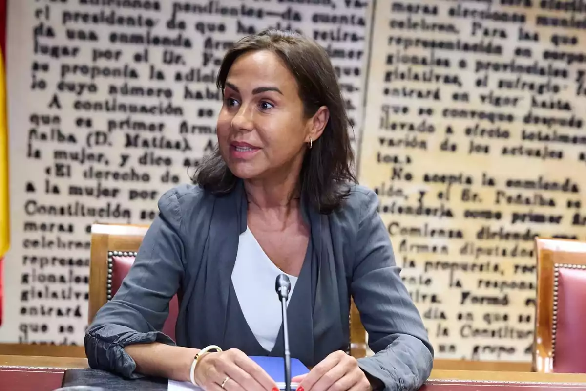 Mujer sentada en una mesa con un micrófono frente a ella y un fondo de texto grande en la pared Mujer sentada en una mesa con un micrófono frente a ella y un fondo de texto grande en la pared