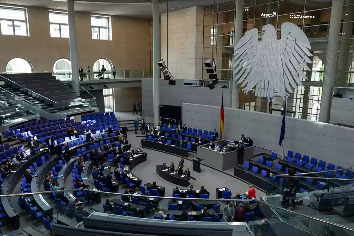 Vista interior del parlamento alemán con varias personas sentadas y un gran águila en la pared Vista interior del parlamento alemán con varias personas sentadas y un gran águila en la pared