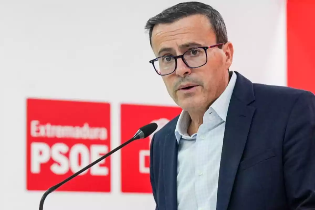 Hombre con gafas y traje hablando frente a un micrófono con carteles de Extremadura y PSOE al fondo Hombre con gafas y traje hablando frente a un micrófono con carteles de Extremadura y PSOE al fondo