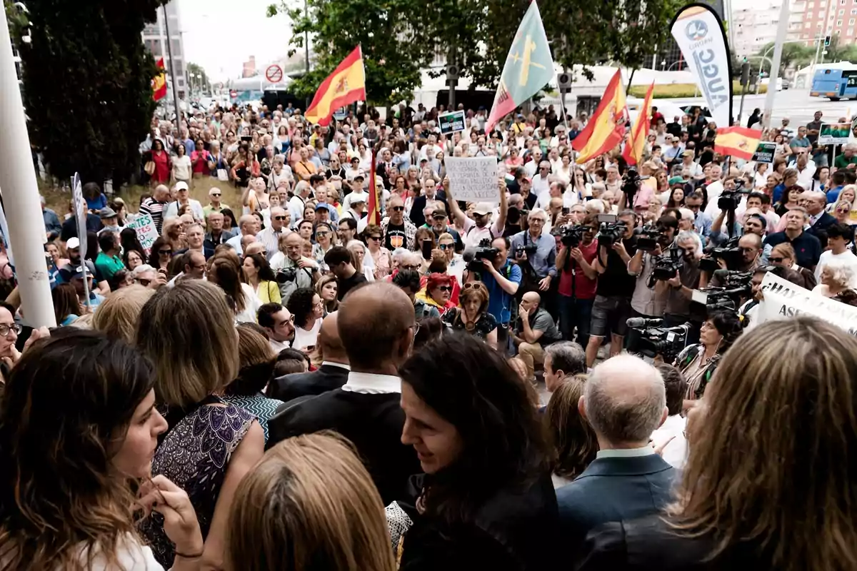 Multitud de personas reunidas en una manifestación al aire libre con banderas de España y pancartas, mientras varios periodistas y cámaras cubren el evento. Multitud de personas reunidas en una manifestación al aire libre con banderas de España y pancartas, mientras varios periodistas y cámaras cubren el evento.