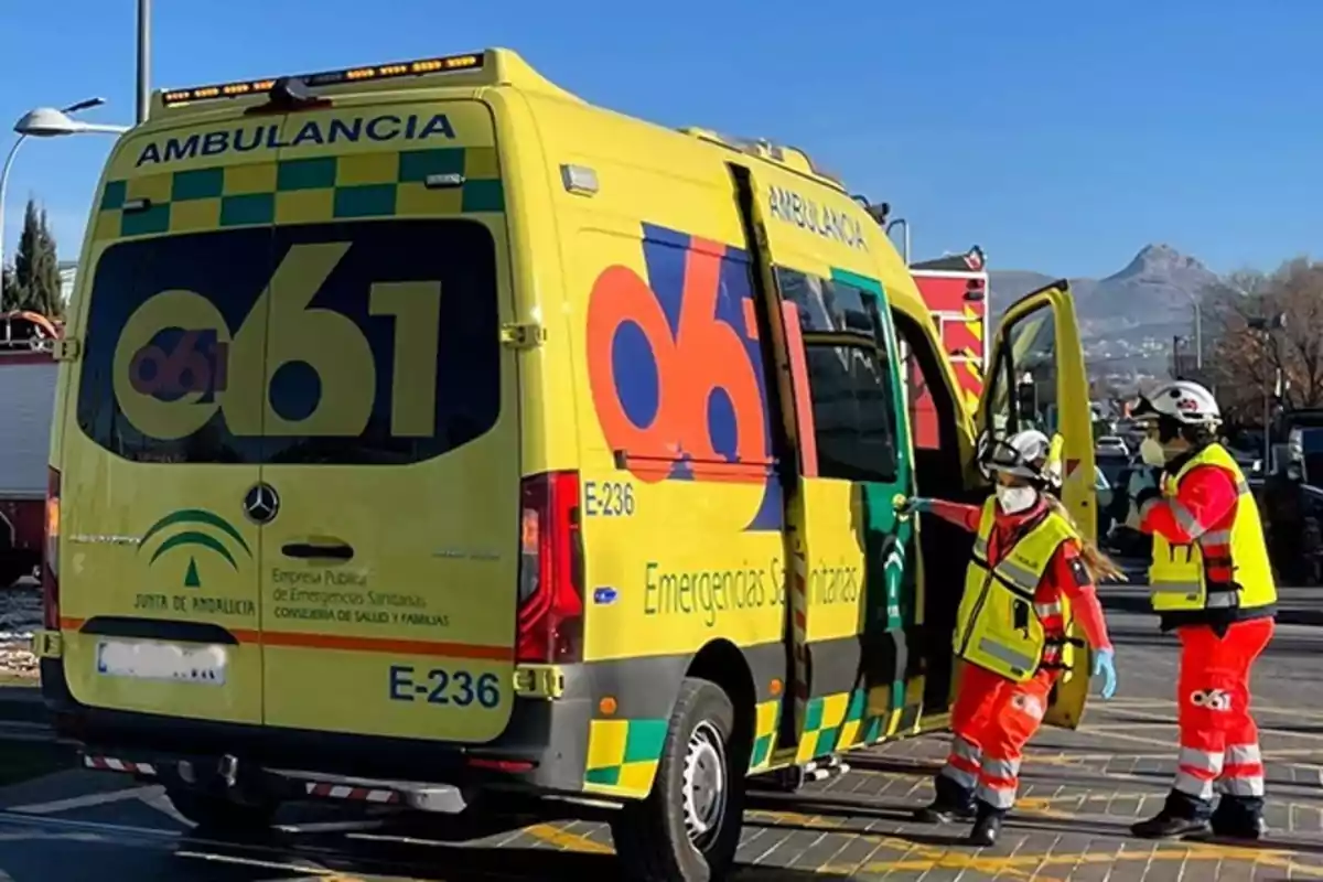 Ambulancia amarilla de emergencias sanitarias de Andalucía estacionada con la puerta lateral abierta y dos trabajadores sanitarios con chalecos reflectantes y cascos junto a ella en un entorno urbano y montañoso. Ambulancia amarilla de emergencias sanitarias de Andalucía estacionada con la puerta lateral abierta y dos trabajadores sanitarios con chalecos reflectantes y cascos junto a ella en un entorno urbano y montañoso.