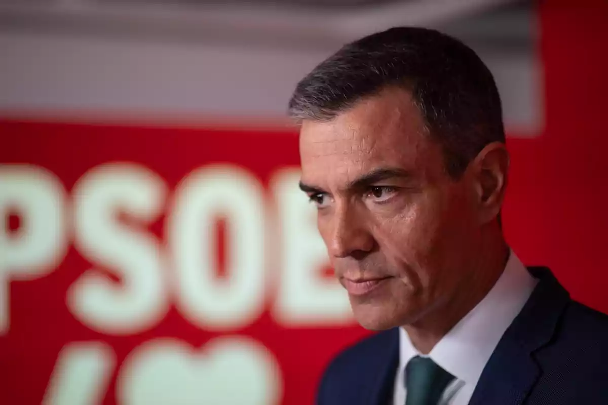 Hombre de cabello corto y canoso con expresión seria frente a un fondo rojo con letras blancas desenfocadas Hombre de cabello corto y canoso con expresión seria frente a un fondo rojo con letras blancas desenfocadas