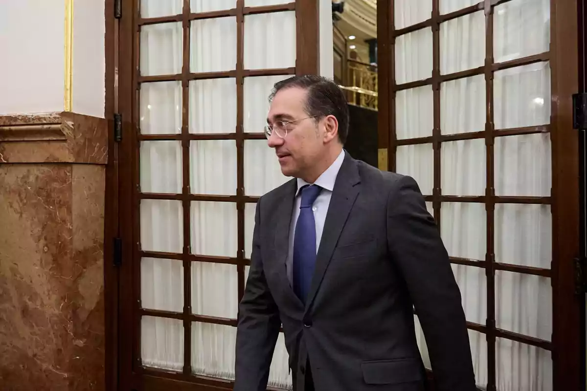 Hombre con gafas y traje oscuro entrando por una puerta de madera con cristales en un edificio elegante Hombre con gafas y traje oscuro entrando por una puerta de madera con cristales en un edificio elegante