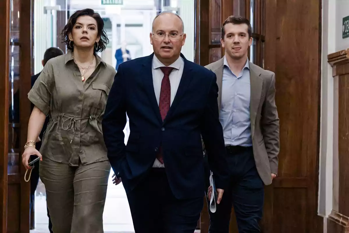 Tres personas caminando por un pasillo elegante, una mujer con mono verde, un hombre con traje azul y corbata roja y otro hombre con saco beige y camisa azul Tres personas caminando por un pasillo elegante, una mujer con mono verde, un hombre con traje azul y corbata roja y otro hombre con saco beige y camisa azul