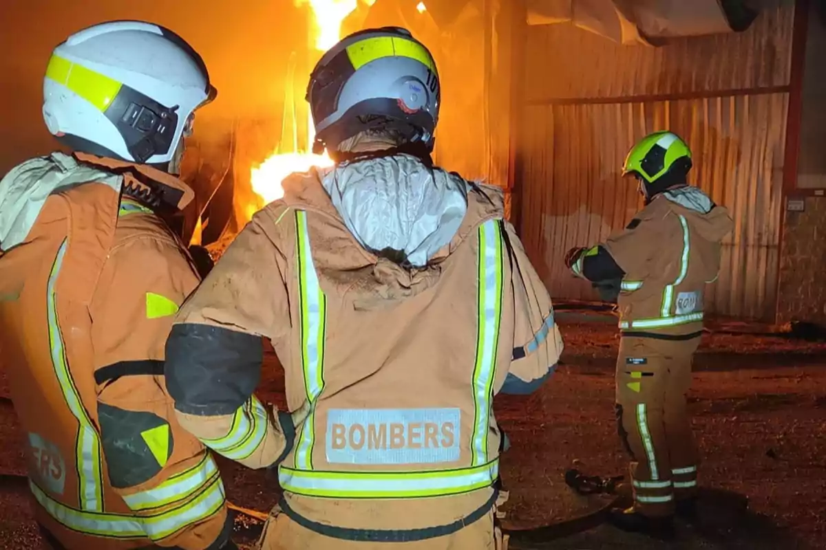 Tres bomberos con trajes y cascos protectores observan un incendio en un edificio industrial durante la noche Tres bomberos con trajes y cascos protectores observan un incendio en un edificio industrial durante la noche