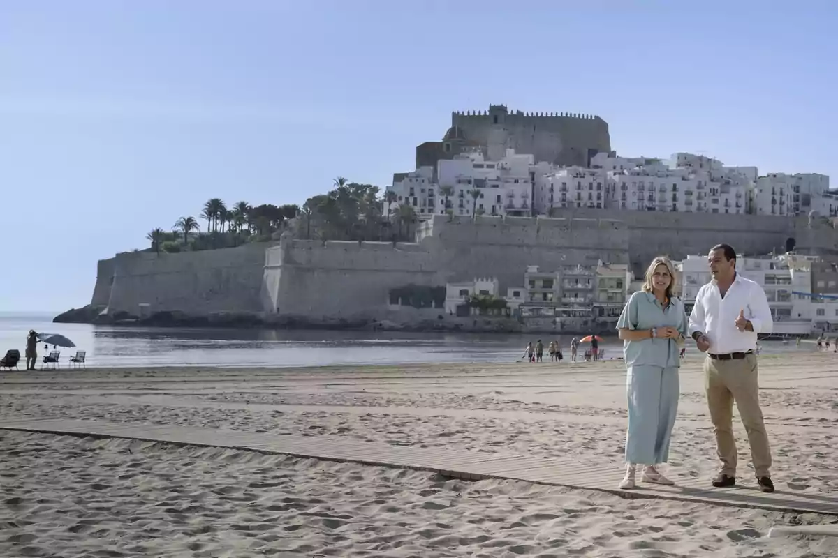 Dos personas conversan en una playa con un castillo y casas blancas al fondo en un día soleado. Dos personas conversan en una playa con un castillo y casas blancas al fondo en un día soleado.