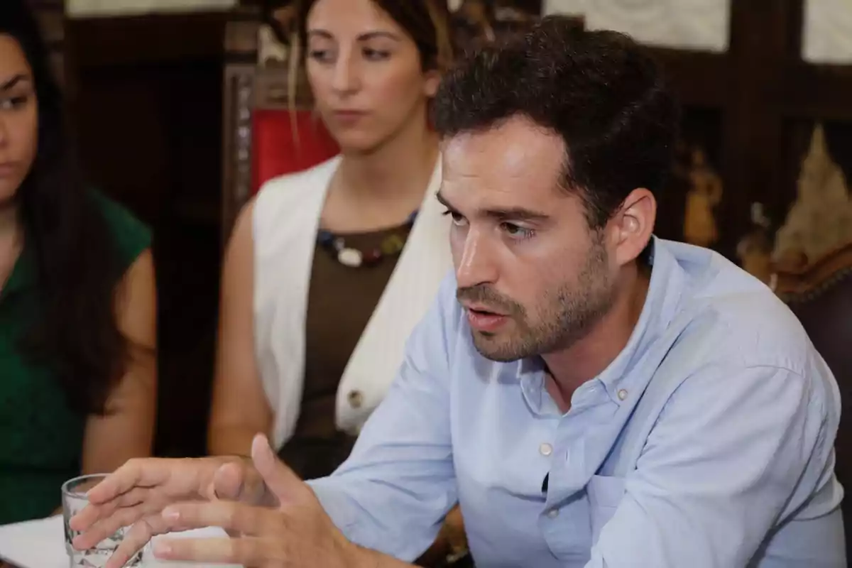 Un hombre con camisa azul claro habla gesticulando con las manos mientras dos mujeres lo escuchan atentamente en una reunión. Un hombre con camisa azul claro habla gesticulando con las manos mientras dos mujeres lo escuchan atentamente en una reunión.