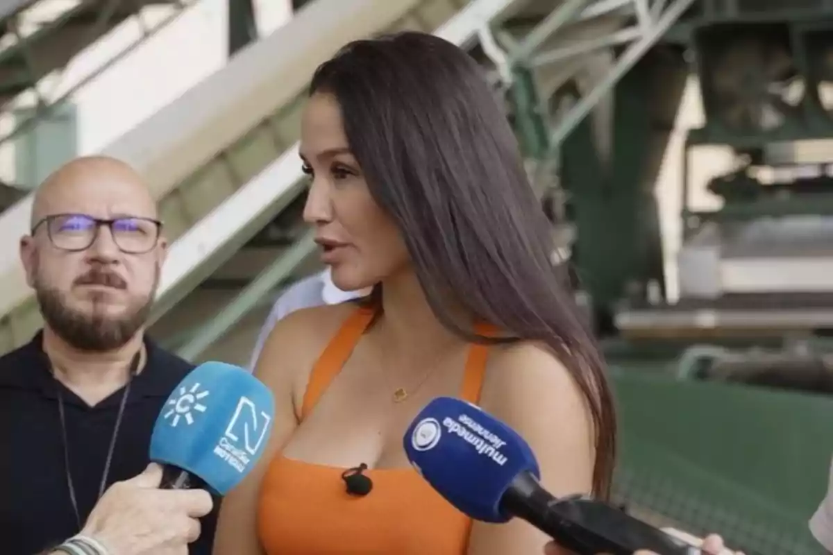 Una mujer con vestido naranja es entrevistada por varios medios de comunicación mientras un hombre con gafas observa en segundo plano en un entorno industrial.