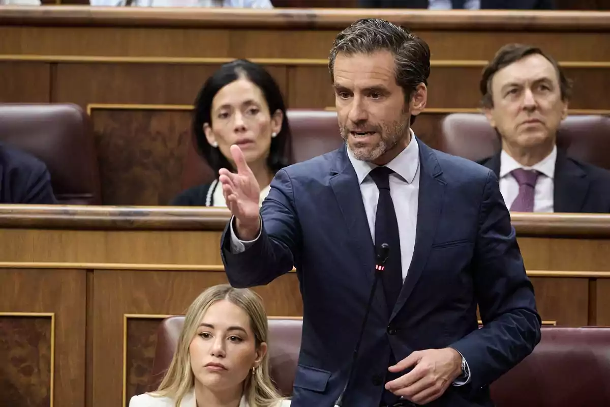 Un hombre de traje azul habla con gesto enfático en un parlamento mientras otras personas lo observan desde sus asientos Un hombre de traje azul habla con gesto enfático en un parlamento mientras otras personas lo observan desde sus asientos