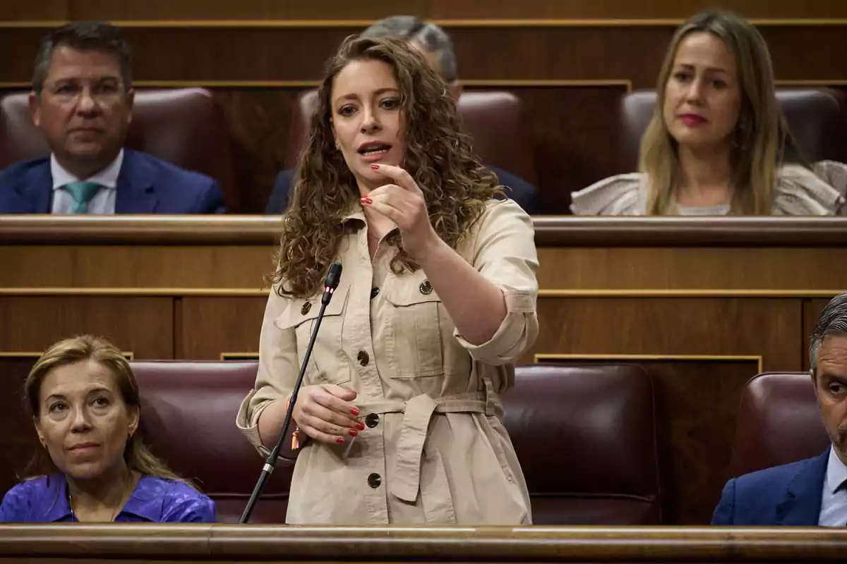 Mujer de cabello rizado hablando de pie frente a un micrófono en un parlamento rodeada de otras personas sentadas