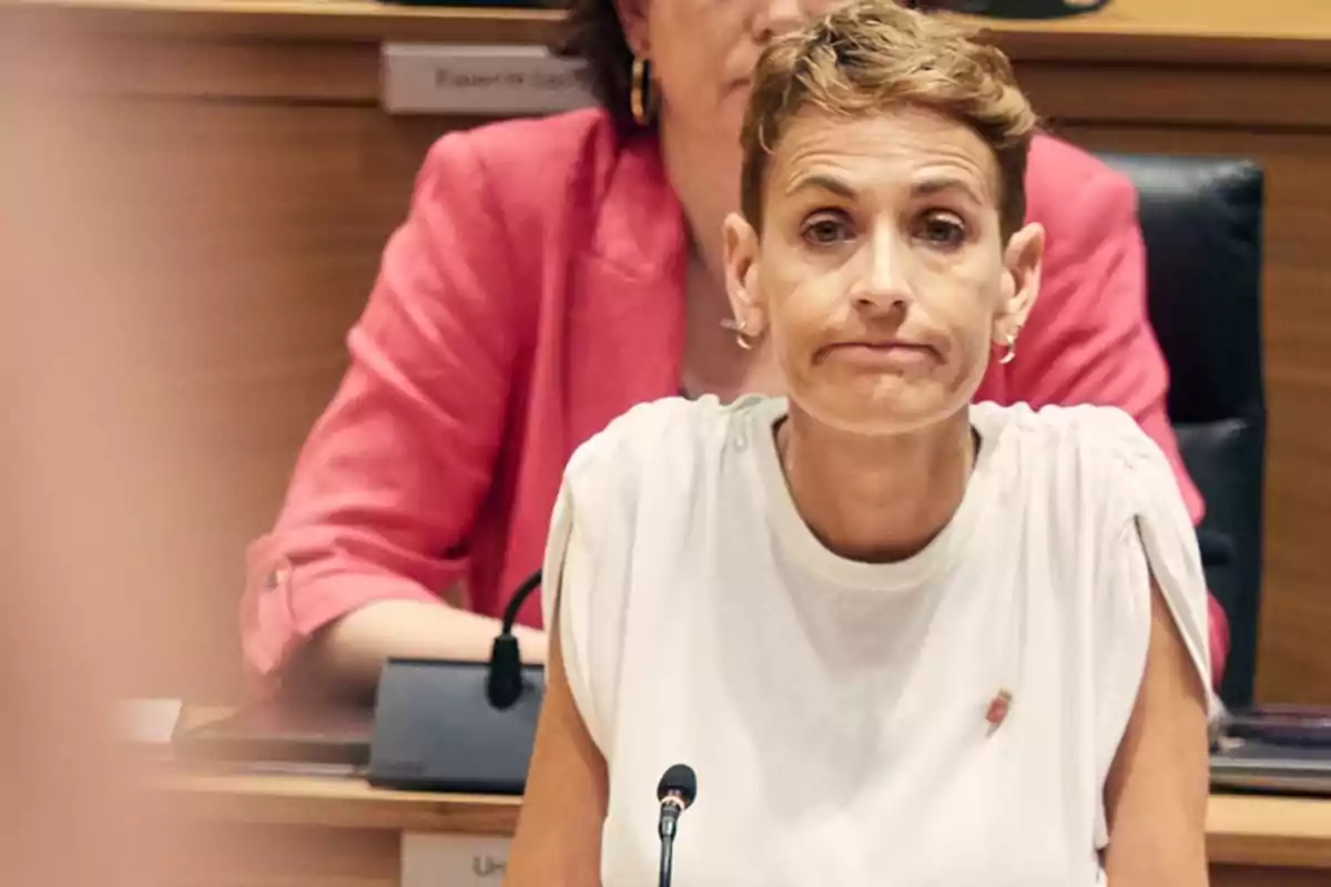 Mujer de cabello corto y rubio con expresión seria sentada frente a un micrófono en una sala de reuniones Mujer de cabello corto y rubio con expresión seria sentada frente a un micrófono en una sala de reuniones