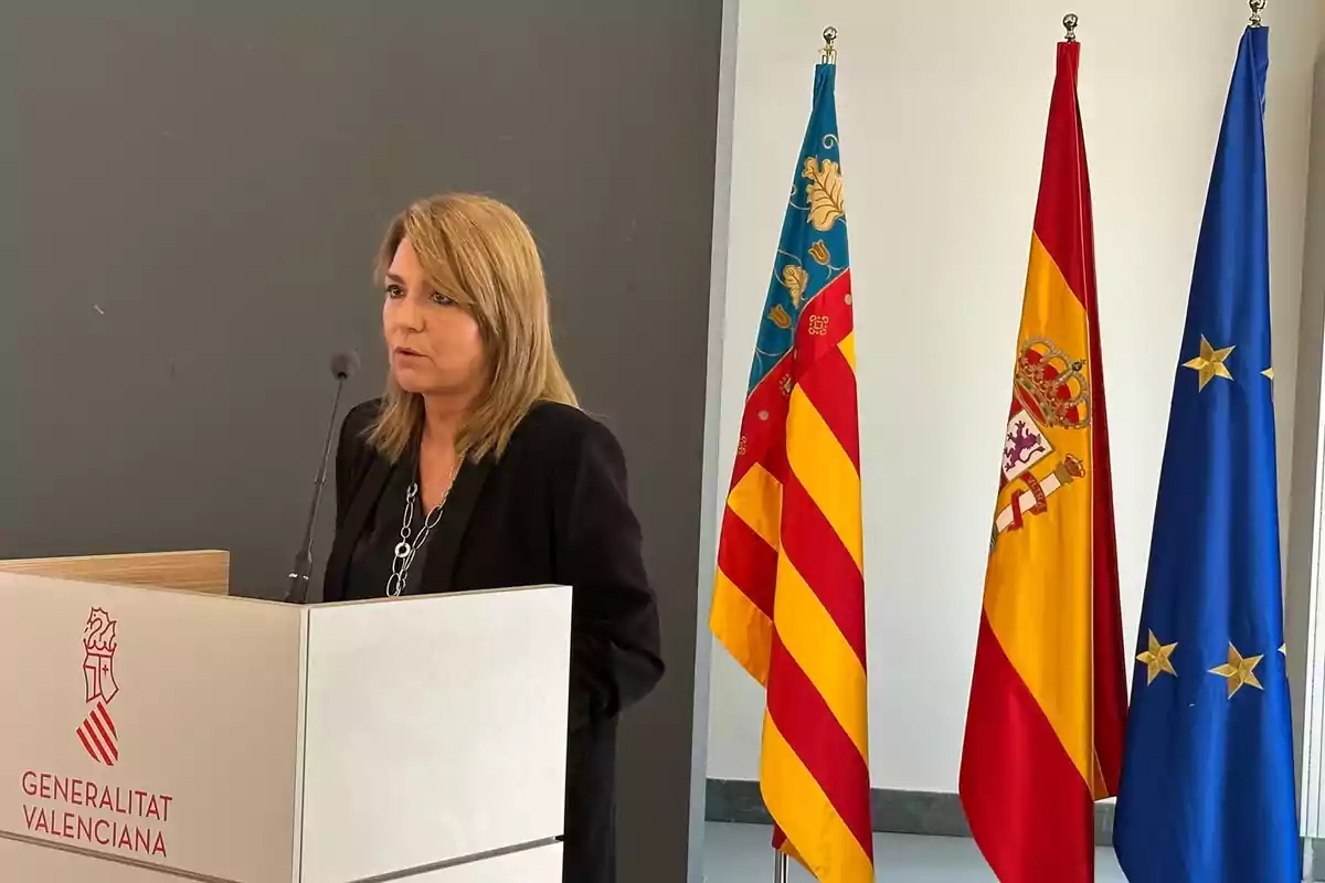 Mujer hablando en un atril de la Generalitat Valenciana con las banderas de la Comunidad Valenciana, España y la Unión Europea al fondo
