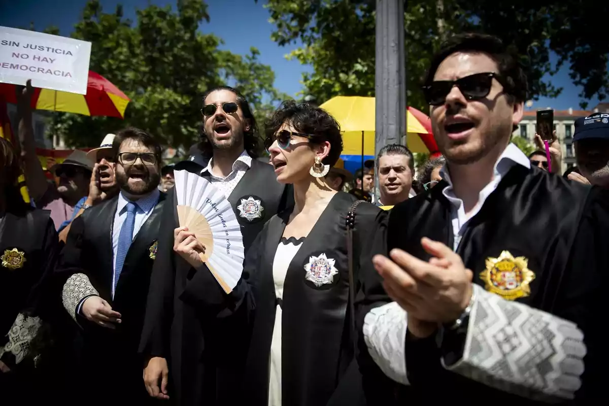 Grupo de personas vestidas con togas negras y gafas de sol participando en una manifestación al aire libre, algunas sostienen abanicos y al fondo se ven pancartas y paraguas de colores.
