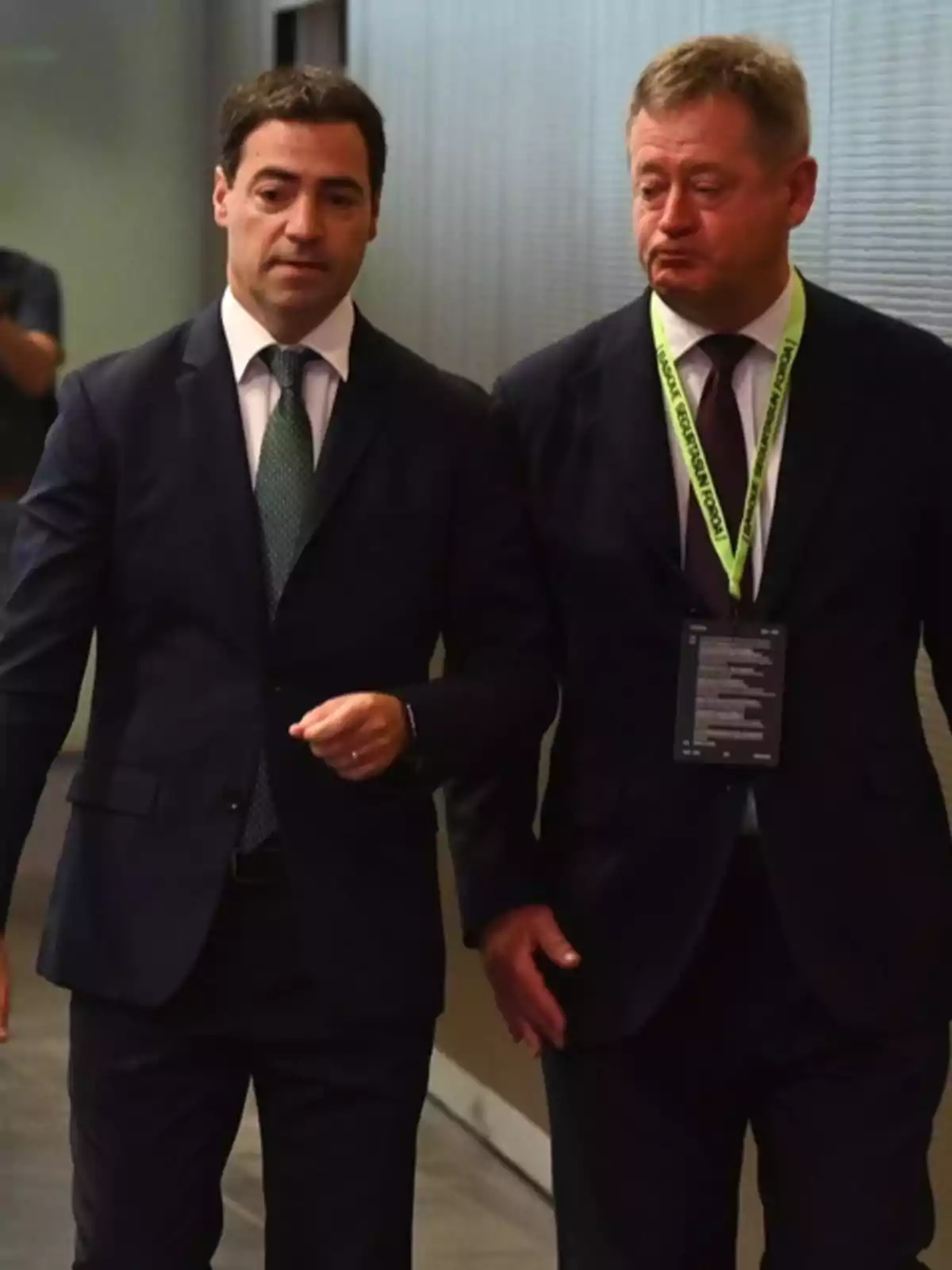 Dos hombres vestidos con traje oscuro caminan juntos en un pasillo mientras conversan, uno de ellos lleva una acreditación colgada al cuello. Dos hombres vestidos con traje oscuro caminan juntos en un pasillo mientras conversan, uno de ellos lleva una acreditación colgada al cuello.