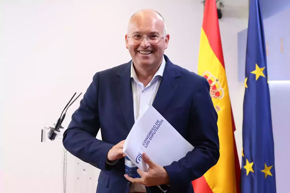 Hombre sonriente con gafas y traje azul sostiene una carpeta y una botella de agua frente a las banderas de España y la Unión Europea
