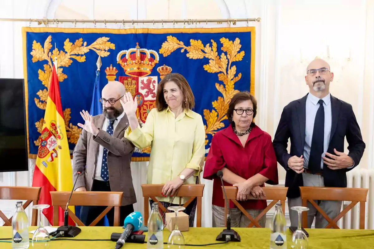 Cuatro personas de pie frente a una mesa con micrófonos y botellas de agua, al fondo una bandera de España y un tapiz azul con escudo oficial Cuatro personas de pie frente a una mesa con micrófonos y botellas de agua, al fondo una bandera de España y un tapiz azul con escudo oficial