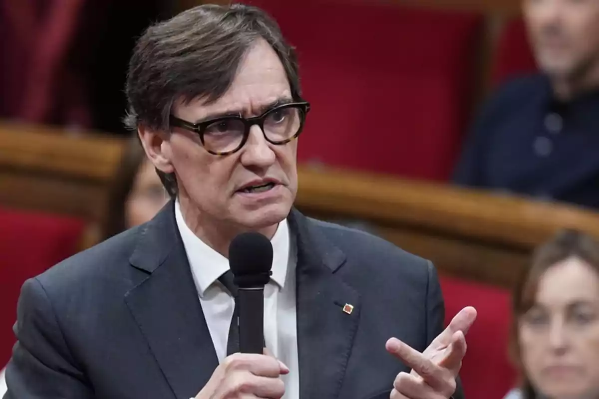 Hombre con gafas y traje oscuro hablando en público mientras sostiene un micrófono Hombre con gafas y traje oscuro hablando en público mientras sostiene un micrófono