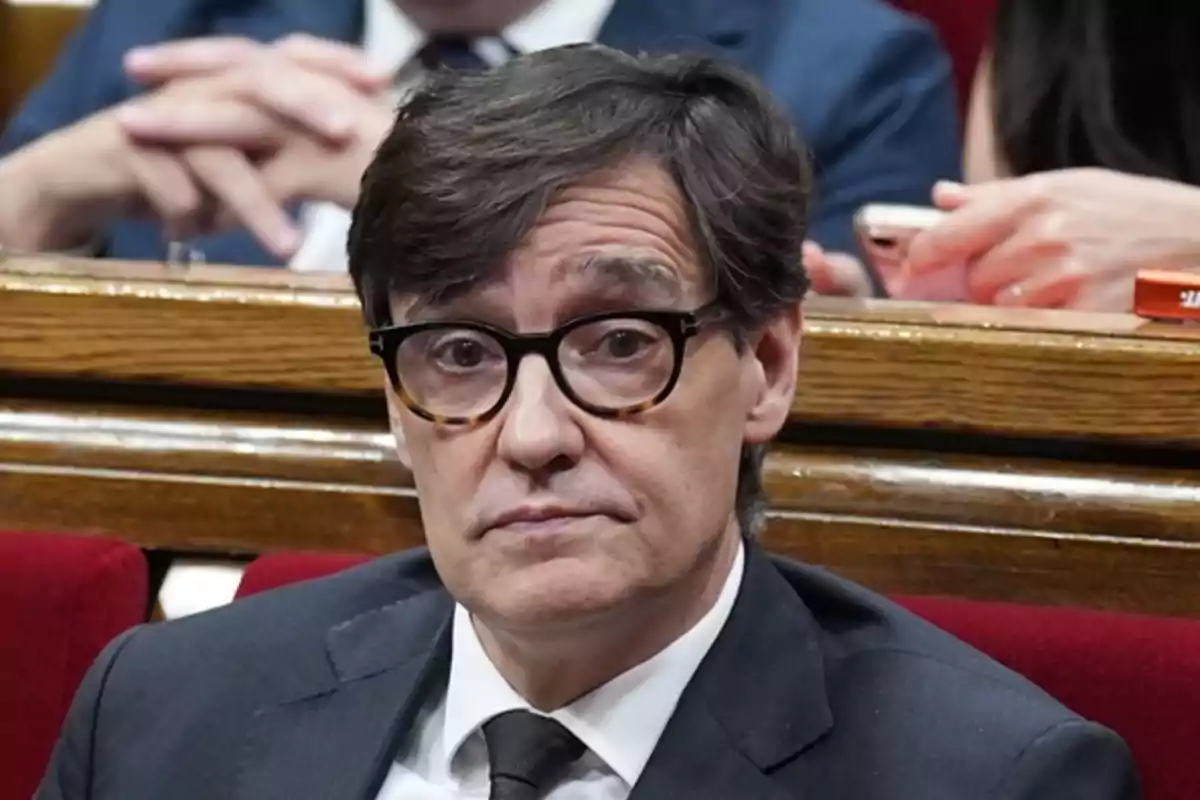 Hombre de cabello oscuro y gafas sentado en un recinto parlamentario con expresión seria