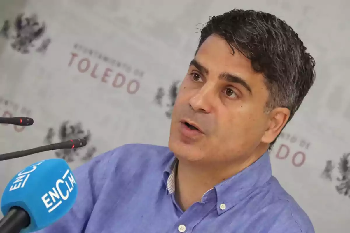 Hombre de cabello corto y camisa azul hablando frente a micrófonos en una rueda de prensa con fondo del Ayuntamiento de Toledo Hombre de cabello corto y camisa azul hablando frente a micrófonos en una rueda de prensa con fondo del Ayuntamiento de Toledo