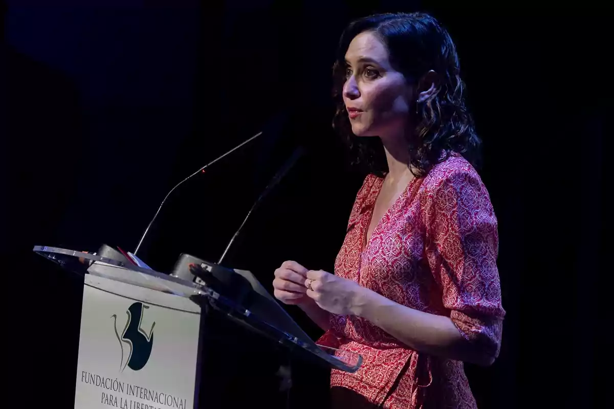 Mujer de cabello ondulado y vestido rojo hablando en un podio con el logo de la Fundación Internacional para la Libertad en un evento iluminado con luz tenue