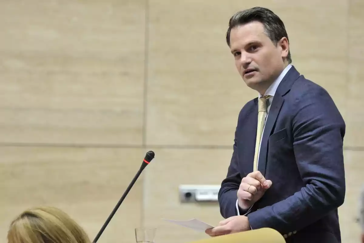 Hombre de traje azul y corbata amarilla hablando frente a un micrófono en una sala de conferencias Hombre de traje azul y corbata amarilla hablando frente a un micrófono en una sala de conferencias