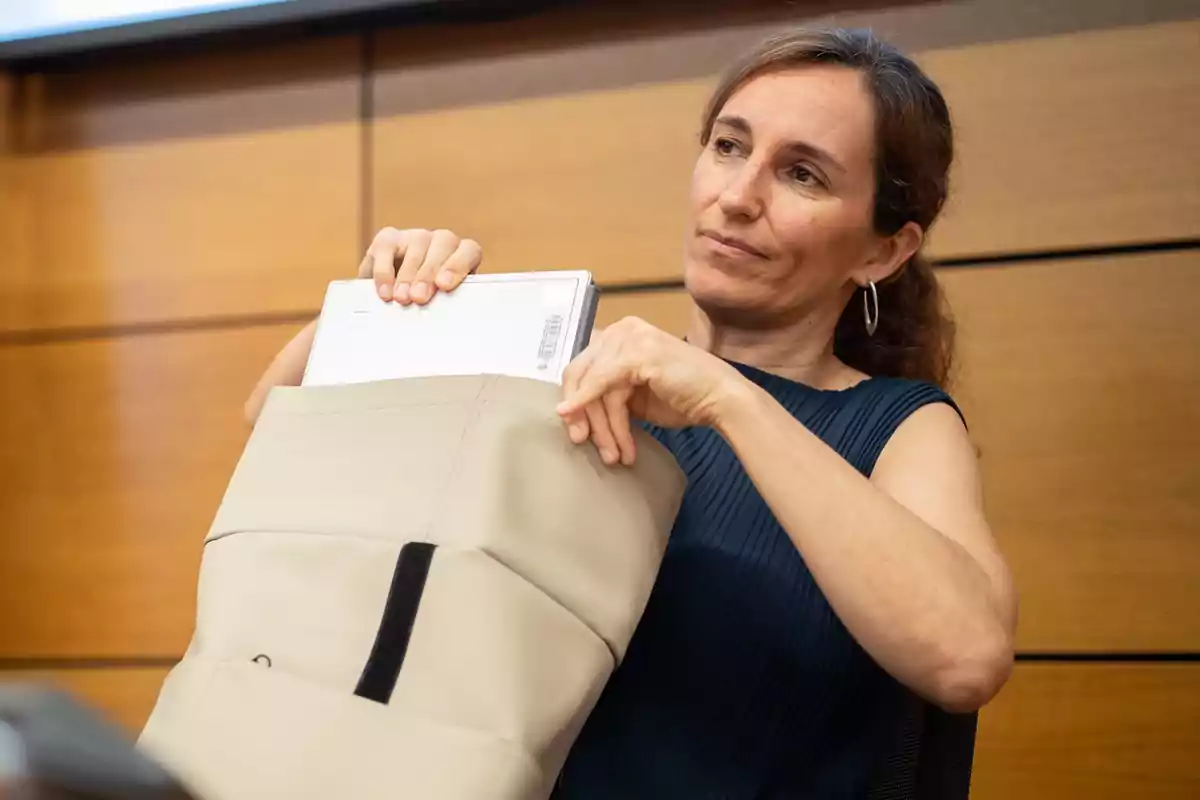 Mujer con blusa sin mangas azul guardando una laptop blanca en una mochila beige frente a una pared de madera