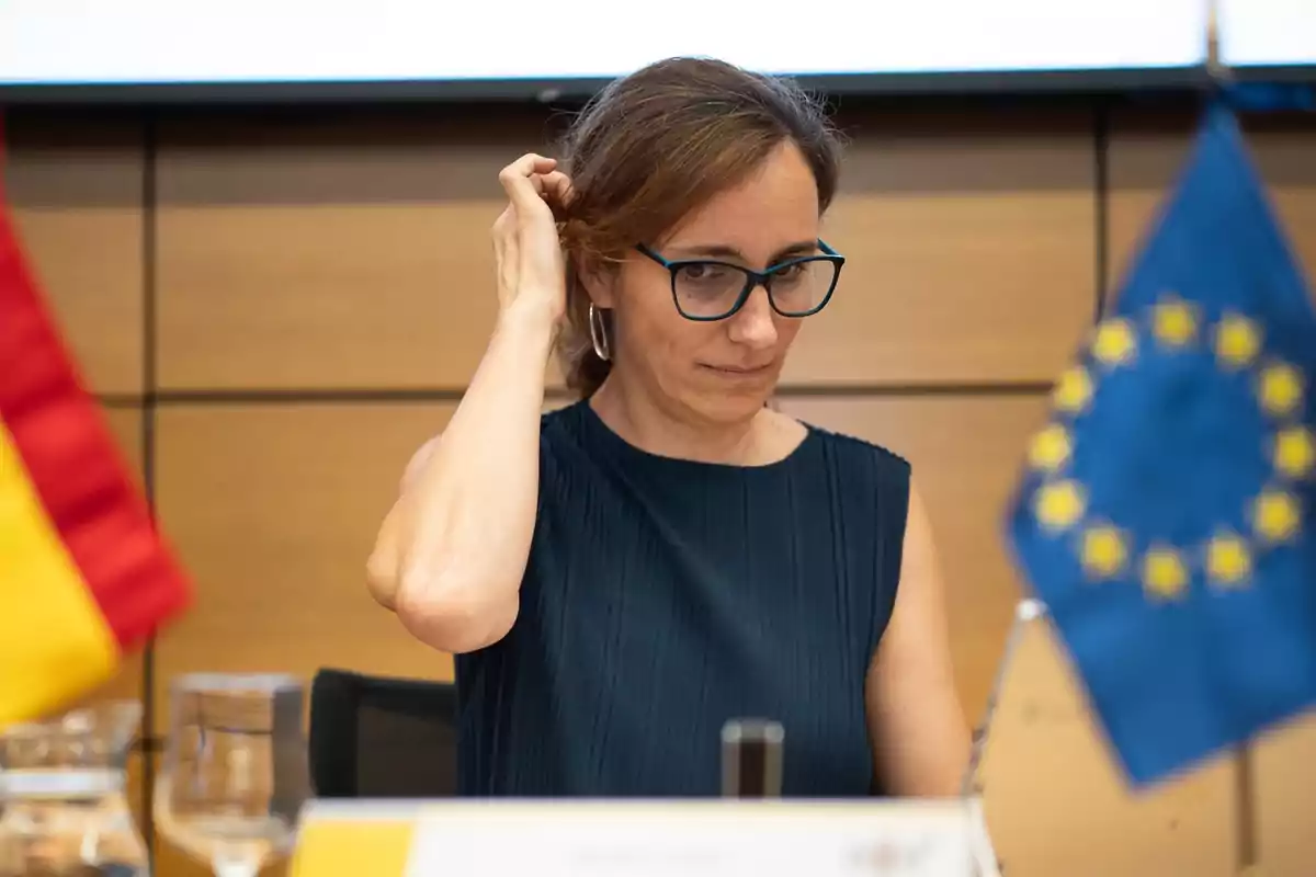 Mujer con gafas y vestido azul oscuro sentada en una mesa entre las banderas de España y la Unión Europea en un entorno de conferencia Mujer con gafas y vestido azul oscuro sentada en una mesa entre las banderas de España y la Unión Europea en un entorno de conferencia