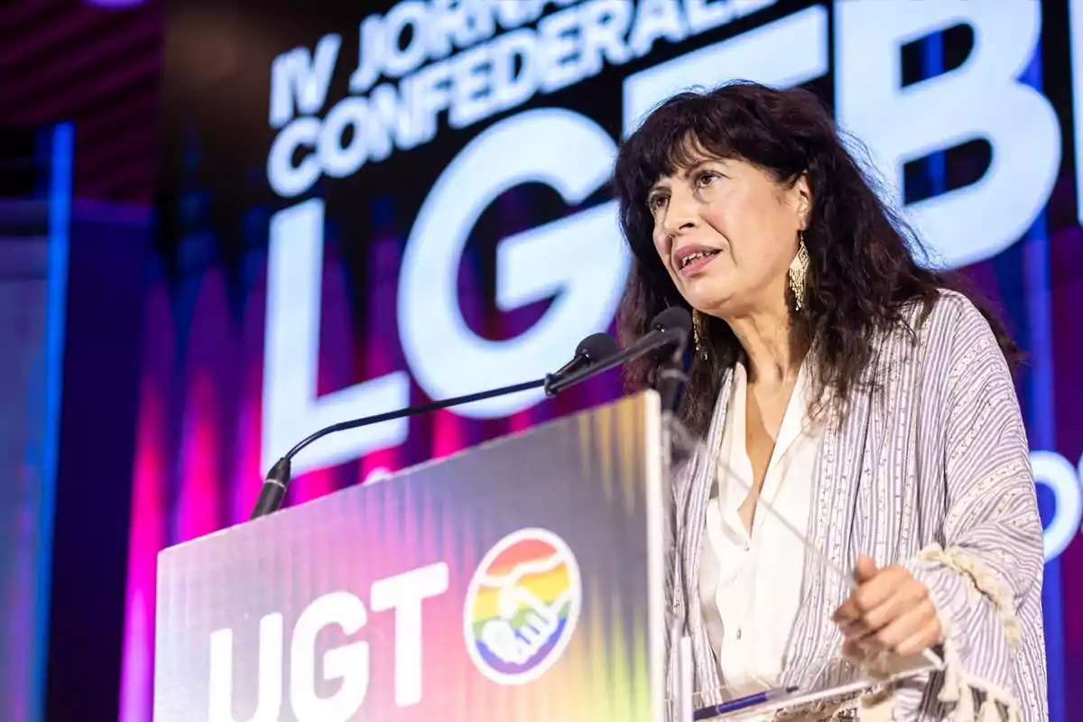 Mujer de cabello oscuro hablando en un podio durante un evento de la UGT con temática LGTB, con fondo colorido y letras grandes en pantalla.