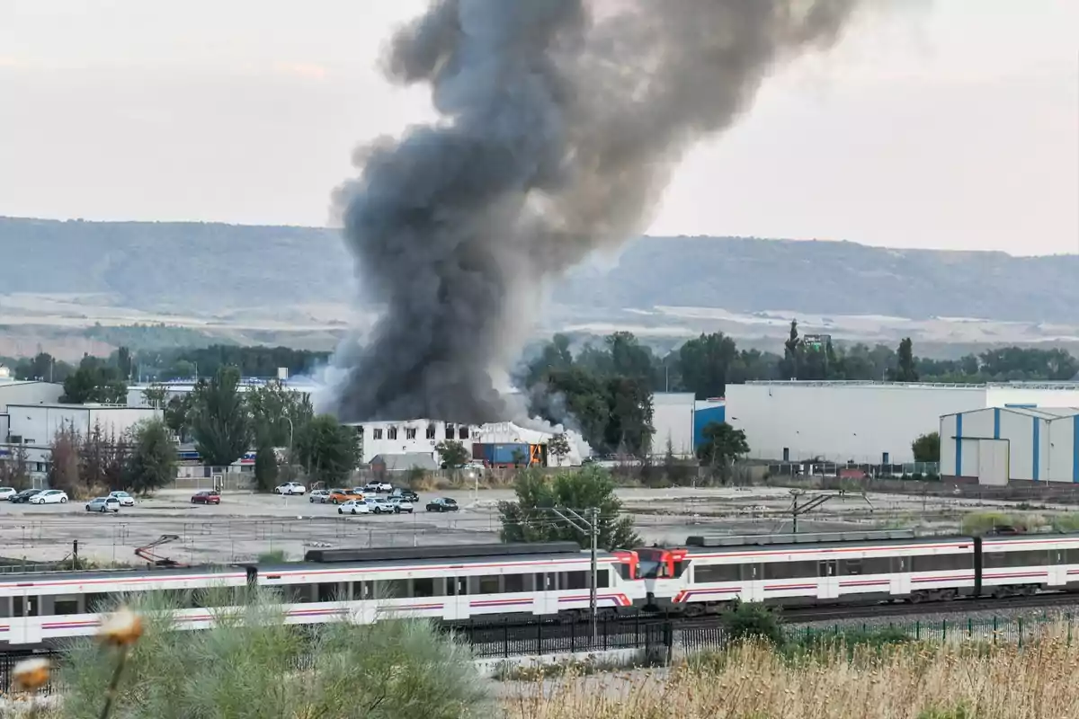 Fábrica en llamas con una gran columna de humo negro y un tren pasando por las vías en primer plano Fábrica en llamas con una gran columna de humo negro y un tren pasando por las vías en primer plano