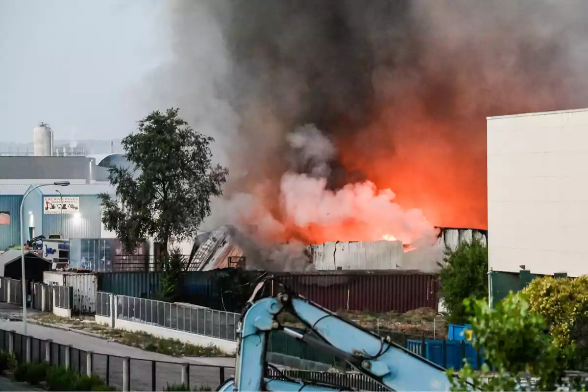 Incendio en una nave industrial con grandes llamas naranjas y humo negro saliendo del edificio mientras maquinaria y árboles se ven en primer plano