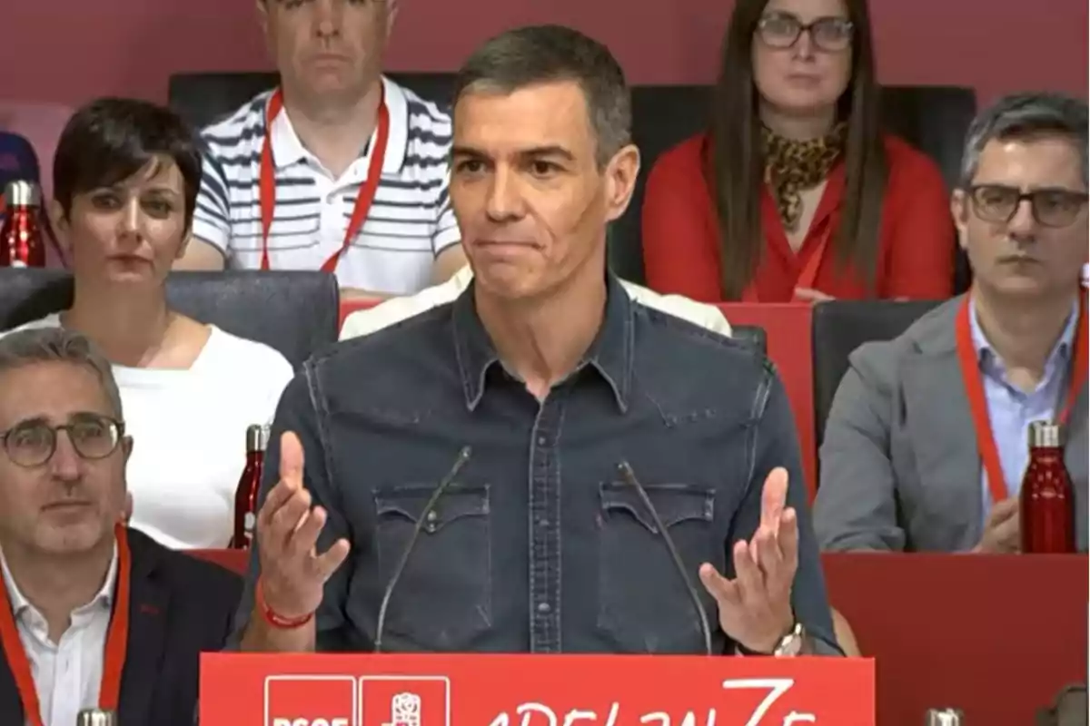 Hombre de cabello corto y canoso hablando en un podio rojo con el logo del PSOE mientras varias personas lo observan sentadas detrás de él
