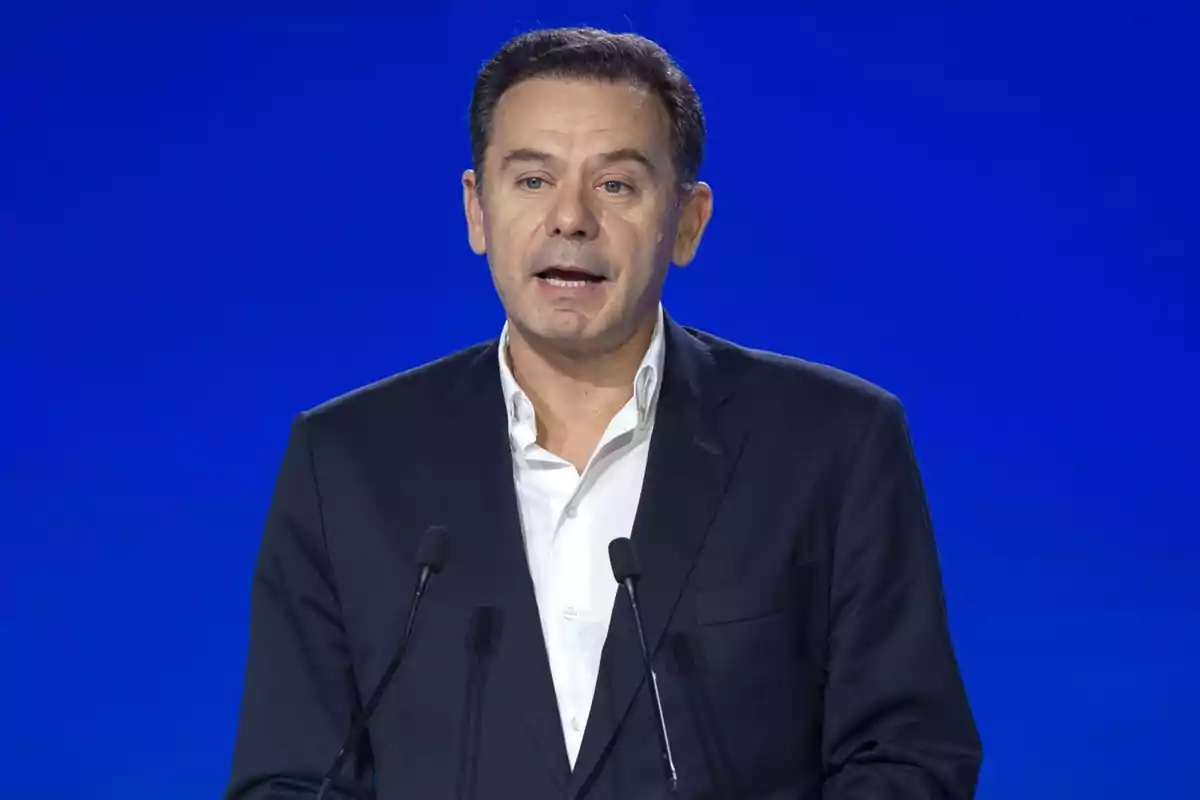 Hombre de cabello corto y oscuro con saco y camisa blanca hablando frente a dos micrófonos sobre un fondo azul Hombre de cabello corto y oscuro con saco y camisa blanca hablando frente a dos micrófonos sobre un fondo azul