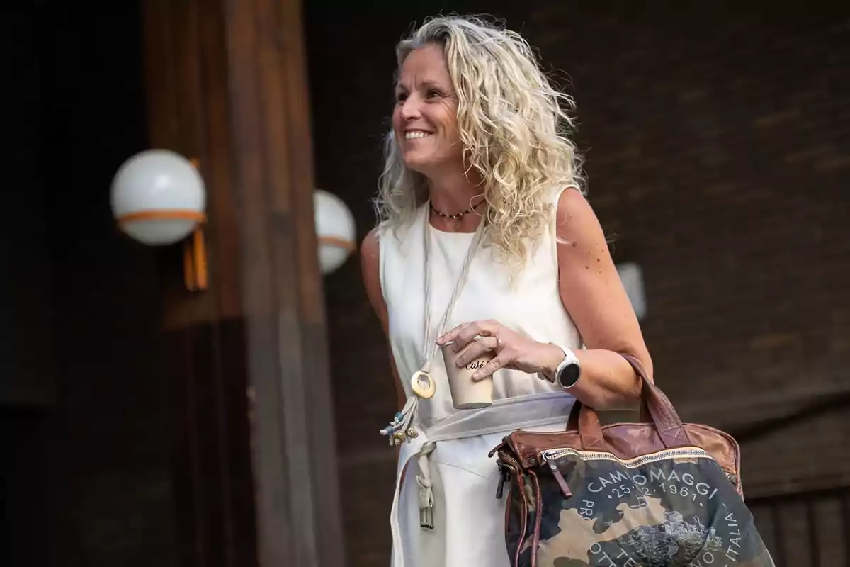Mujer rubia de cabello rizado sonríe mientras sostiene un café y una bolsa grande en un entorno urbano Mujer rubia de cabello rizado sonríe mientras sostiene un café y una bolsa grande en un entorno urbano