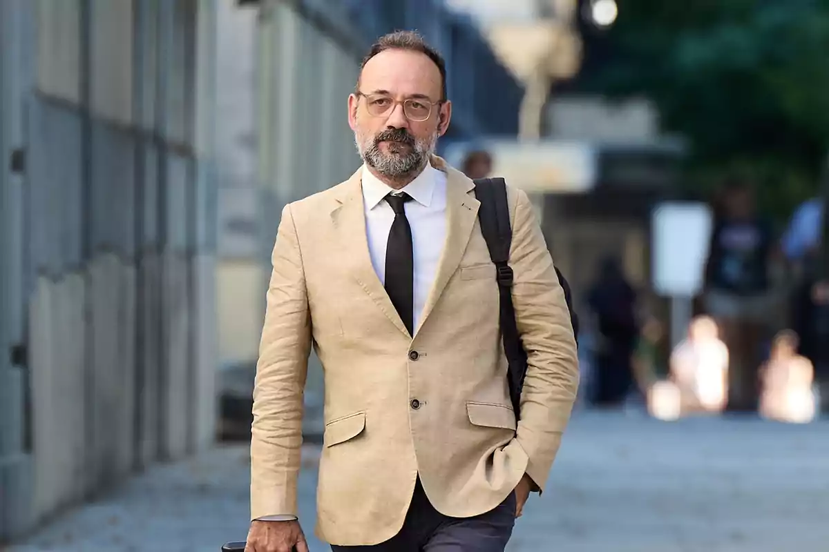 Hombre con barba y gafas camina por la calle vistiendo saco beige, camisa blanca y corbata negra mientras lleva una mochila negra al hombro Hombre con barba y gafas camina por la calle vistiendo saco beige, camisa blanca y corbata negra mientras lleva una mochila negra al hombro