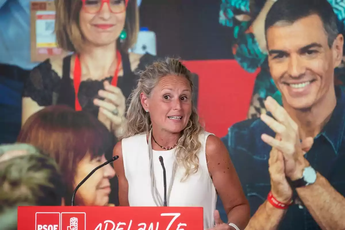 Mujer hablando en un atril rojo con el logo del PSOE y un fondo con personas aplaudiendo Mujer hablando en un atril rojo con el logo del PSOE y un fondo con personas aplaudiendo