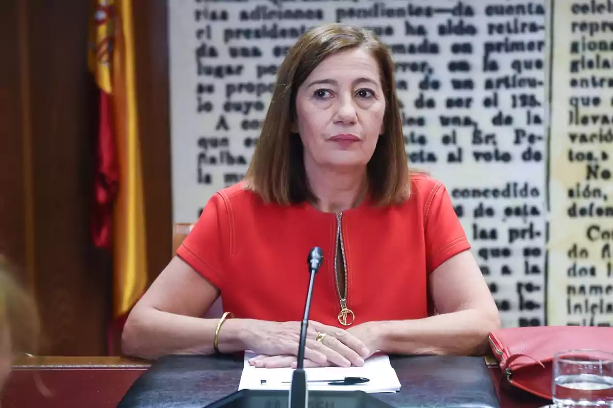 Mujer de cabello castaño y liso sentada en una mesa con micrófono, vistiendo blusa roja y con expresión seria, al fondo una bandera de España y un mural con texto desenfocado.