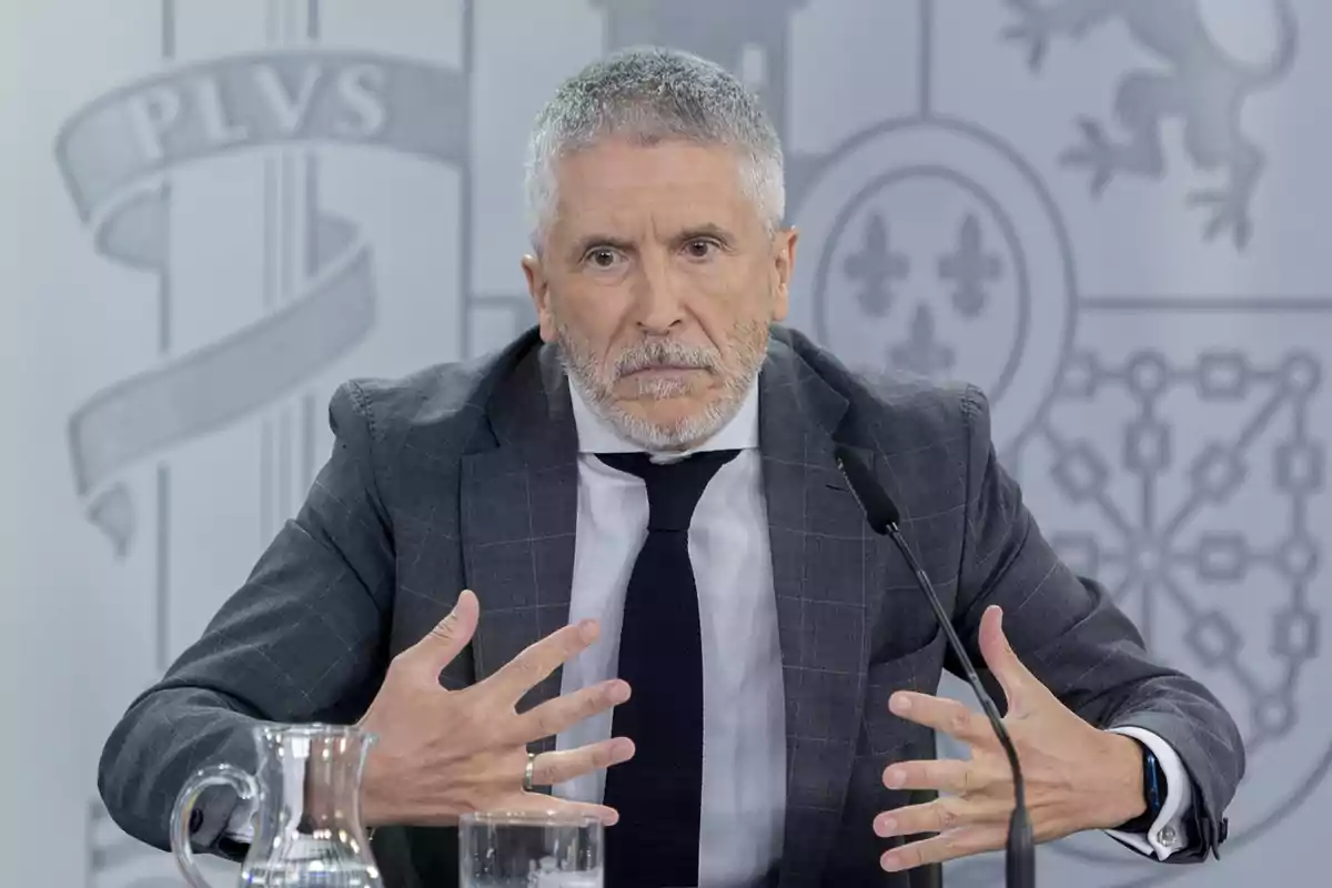 Hombre de cabello canoso y barba sentado en una mesa con micrófono y jarra de agua gesticulando con las manos mientras habla en una conferencia de prensa